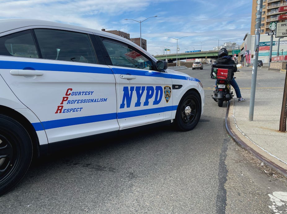 A blue and white NYPD police car is parked behind a motor scooter driver who is at a standstill.