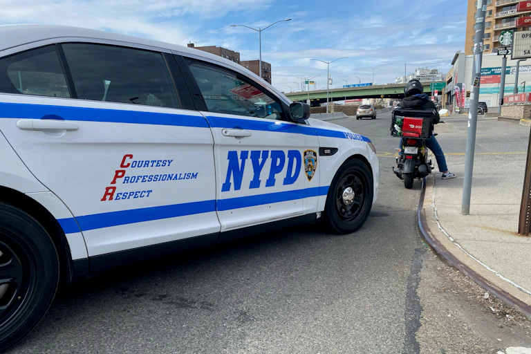 A blue and white NYPD police car is parked behind a motor scooter driver who is at a standstill.