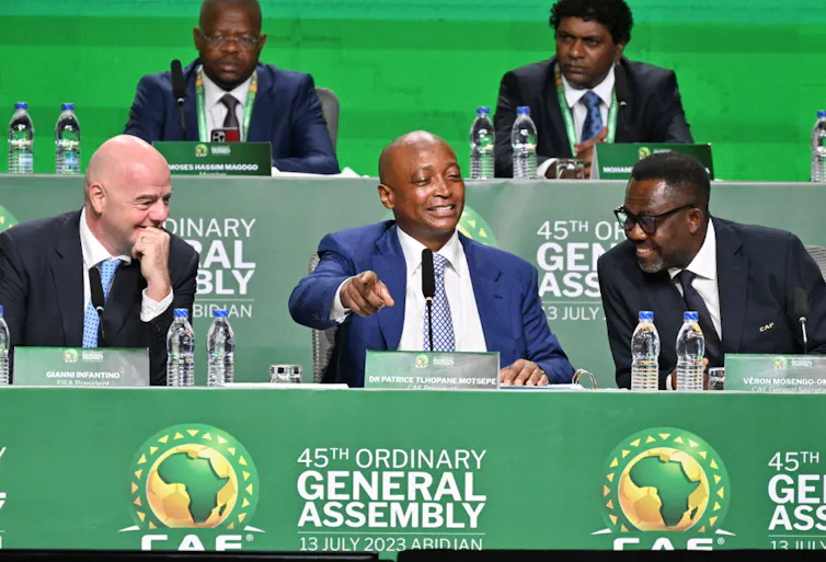Five men in suits sit at tables with microphones and bottles of water in front of them, against a green backdrop.