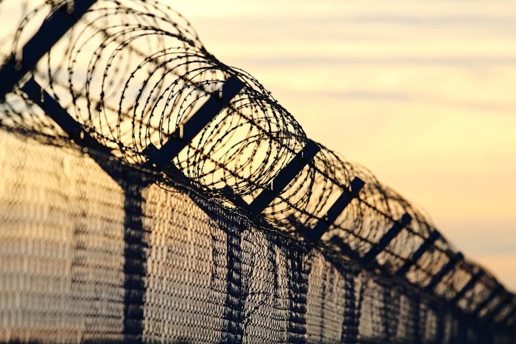 Silhouette of a fence topped with barbed wire, against a yellow sky at dusk