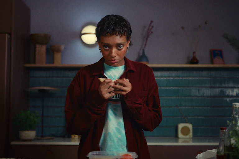 A young black woman looks to camera. Her hair is cropped and she is holding a sandwich.