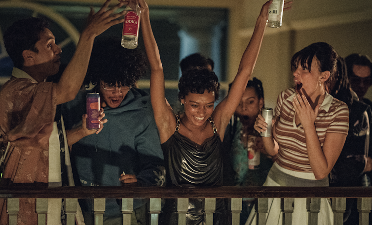 A young woman throws her arms up at a party, a bottle of spirits in each hand.