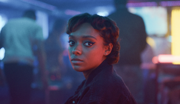 A young black woman with cropped hair and dark eyeliner lit by blue and pink lights.