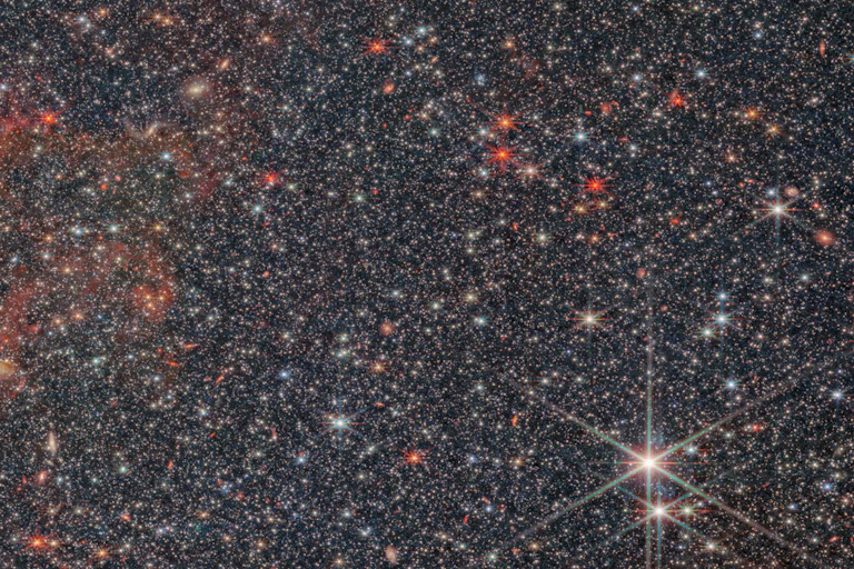 Image of galaxy NGC 6822 captured by Webb’s Near-Infrared Camera, or NIRCam. It shows a densely packed field of stars, mixed with swirls of red gas, on a black background. The stars come in a variety of shapes, sizes and colors. Most are white, but some are yellow or red. The smaller stars appear as dots, but larger stars have distinctive snowflake-like patterns. Two particularly prominent stars are located in the bottom right corner of the image.
