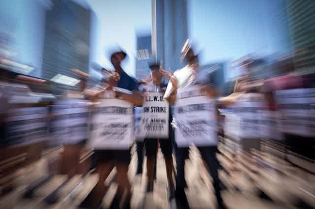 A blurred photo shows striking workers on a sunny day in front of tall buildings.
