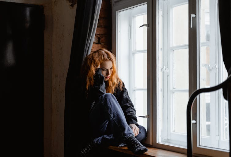Woman with ginger hair sitting by a window looking sad.