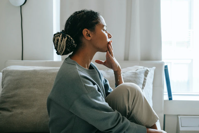Woman with braids sits on a bed looking contemplative.