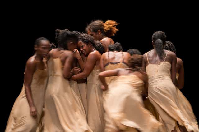 A group of black women dancers in a huddle, wearing nude slip dresses.