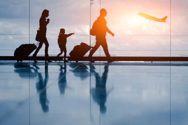 Silhouettes of a family walking along in an airport with a plane taking off in the background. 