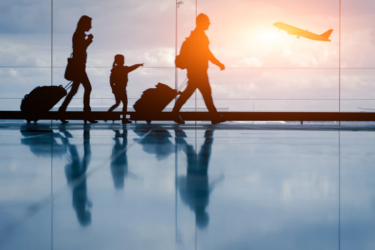 Silhouettes of a family walking along in an airport with a plane taking off in the background.