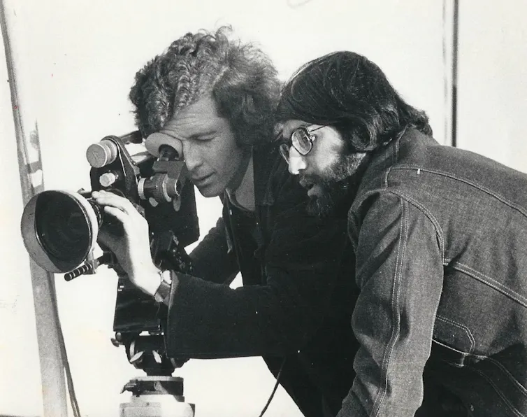 Two men looking down the lens of a film camera in the 1970s.