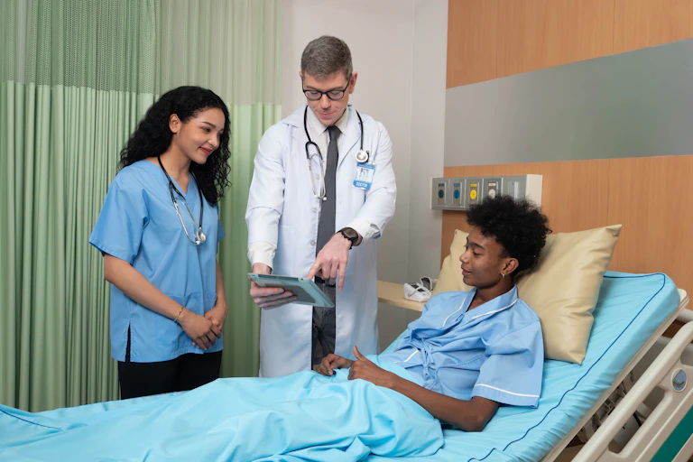 A patient in a hospital bed with a man in a white coat and a younger woman in scrubs