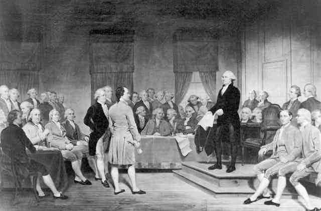 A black and white painting shows George Washington and other men standing around a table together. 