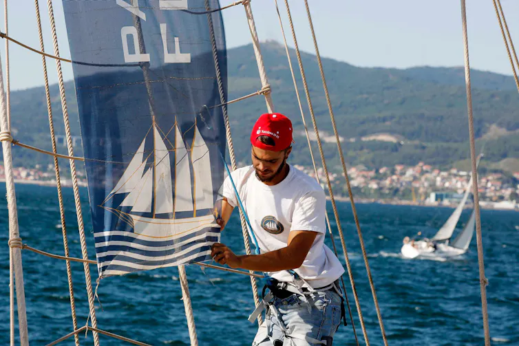 A sailor in a red cap fixes rigging on a sailing boat at sea.