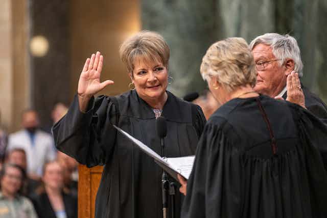 A light-haired woman raising her right hand, wearing a black robe, listens to another person facing her, also wearing a black robe.