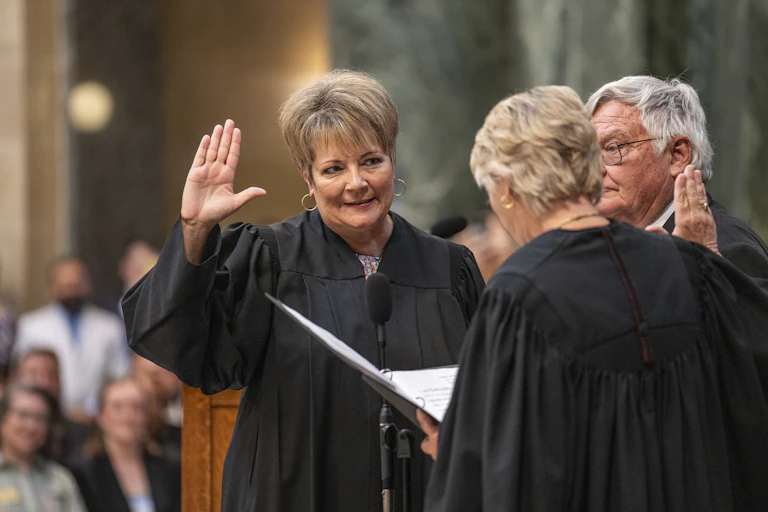 A light-haired woman raising her right hand, wearing a black robe, listens to another person facing her, also wearing a black robe.