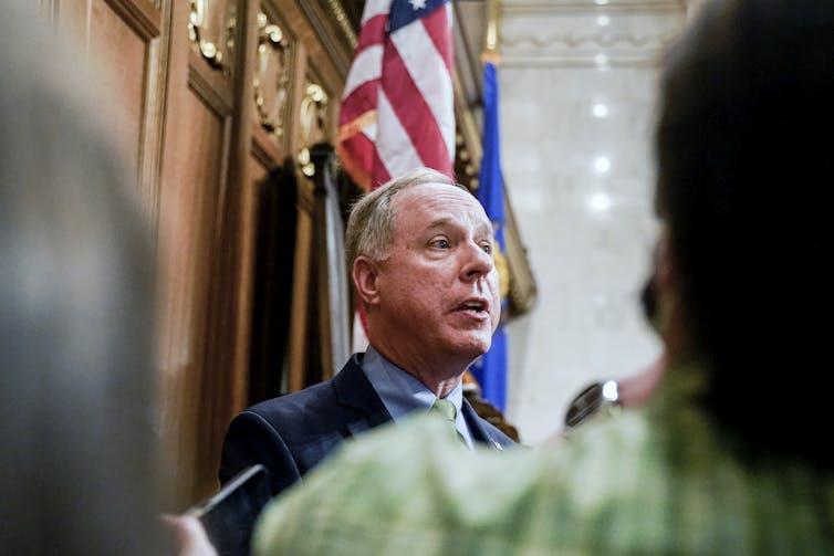 A man in a blue blazer, blue shirt and tie speaking in front of a flag.