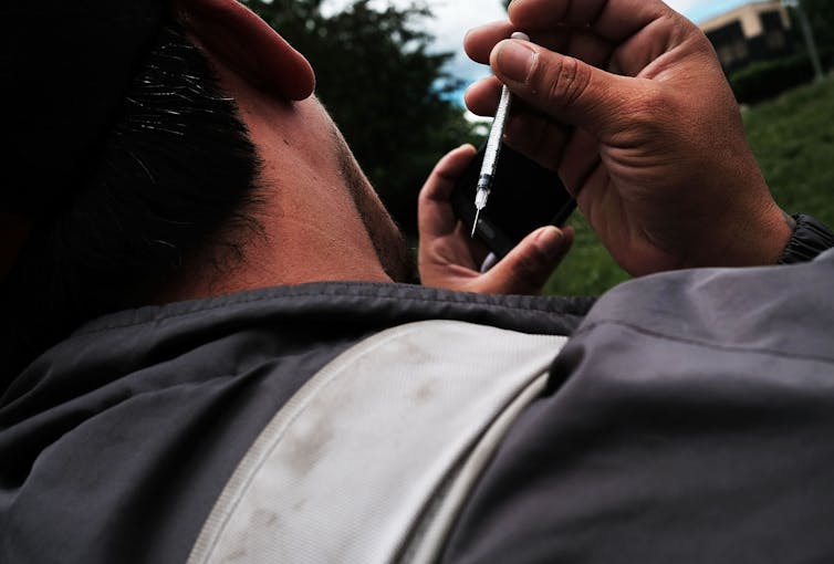 A man with his back to the camera points a heroin needle in the direction of his neck.