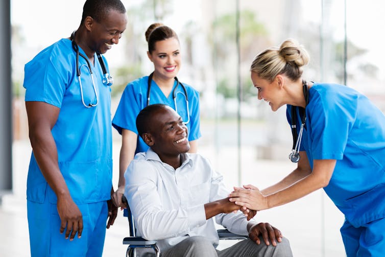 A woman in scrubs shakes hands with a man using a wheelchair in front of two other people in scrubs