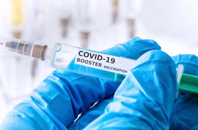 A blue-gloved hand holds up a COVID-19 booster shot syringe.