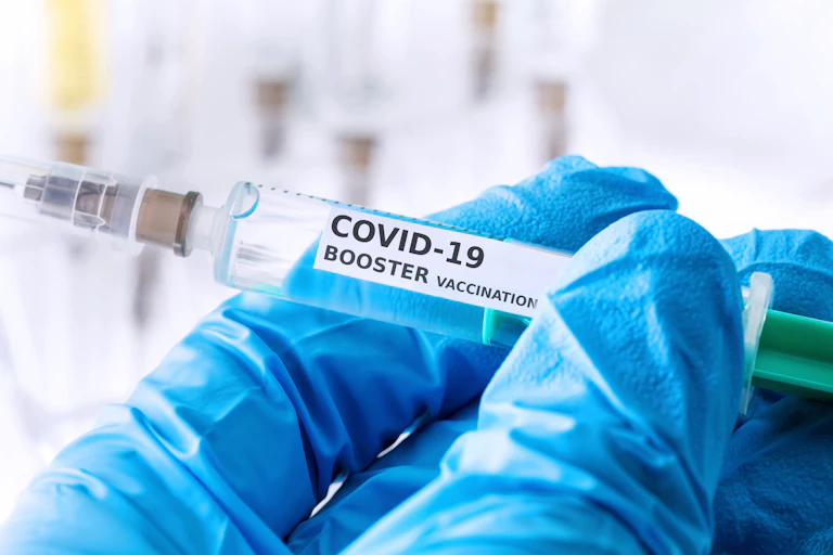 A blue-gloved hand holds up a COVID-19 booster shot syringe.
