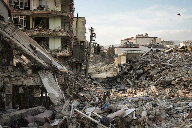 An apartment building leans, its walls are gone and furniture lies under the rubble outside. Other buildings are in similar shape. A person walks on the street among them.