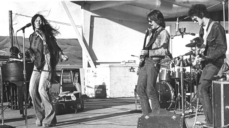 A woman seen on the left at a microphone performing, at right are two men on guitar and all are on a stage.