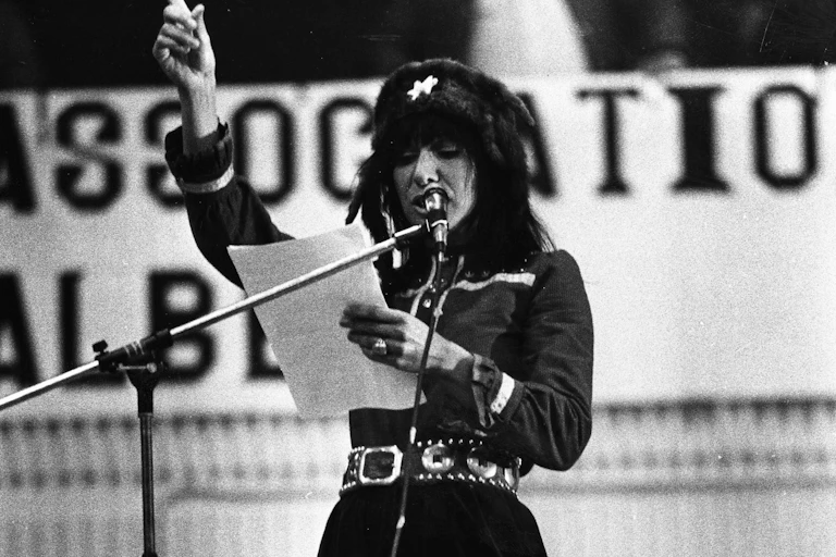 A woman seen with hand raised speaking at microphone.