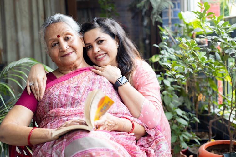 A young Indian women places her arms around an older woman wearing a sari and holding a book.