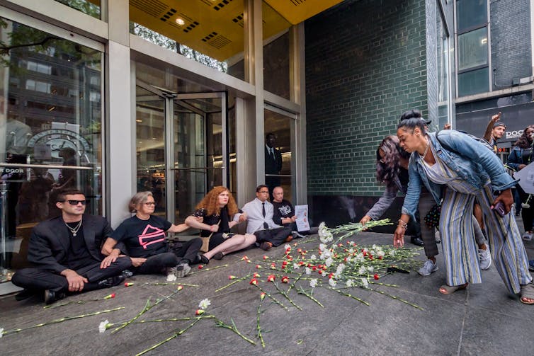 Two women drop flowers on the pavement as five people sit with their arms locked and their backs against a building entrance.