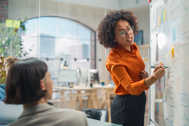 A woman writes on a whiteboard while speaking to a table full of people