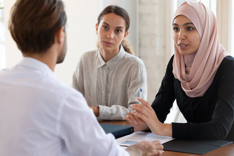 Two women, one wearing a hijab, speak to a man at a table