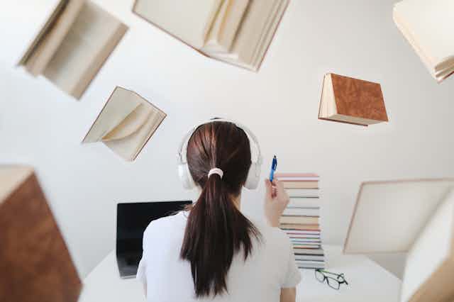 Imagem mostra uma jovem usando headphones lendo livros e estudando em um computador