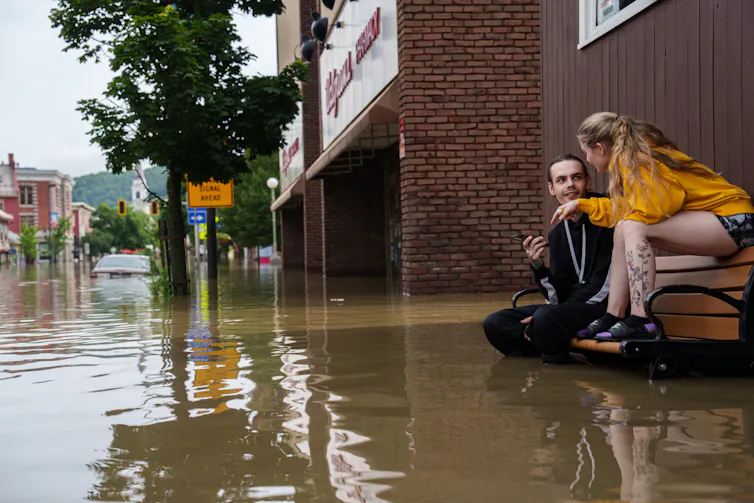Un hombre y una mujer sentados en un banco de un parque con el agua hasta las rodillas del hombre. La mujer está sentada en el respaldo de la silla. Un coche en la calle está inundado hasta el techo.
