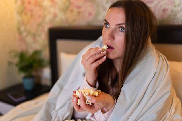 Woman sitting on bed, wrapped in blanket, eating popcorn, watching movie