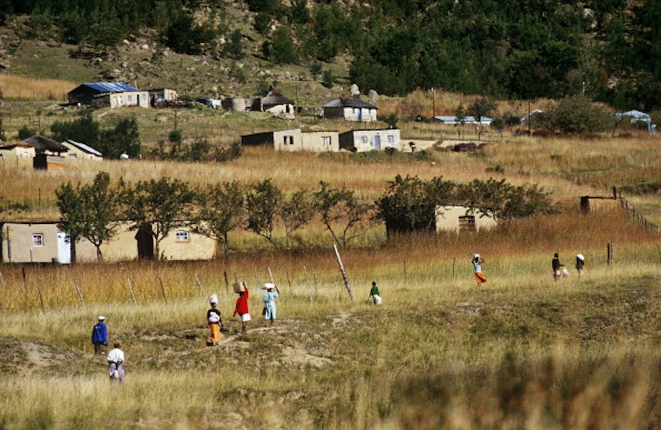 People walk past a field and houses in a rural setting.