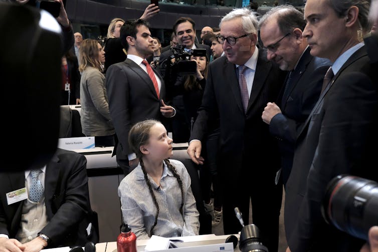Greta Thunberg, ativista climática sueca de 16 anos, participa no evento do Comité Económico e Social Europeu. Sentada, ela está cercada por adultos que estão de pé