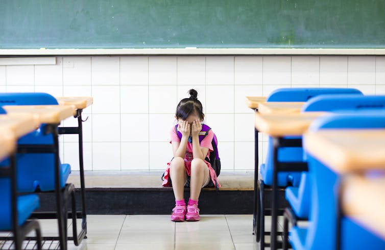 Una niña sola en un salón de clases, con la cabeza entre las manos.