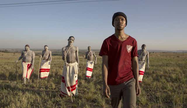 Across an open field, a young man in civilian clothing walks ahead of a group of boys wearing traditional red and white cloths around their waists.