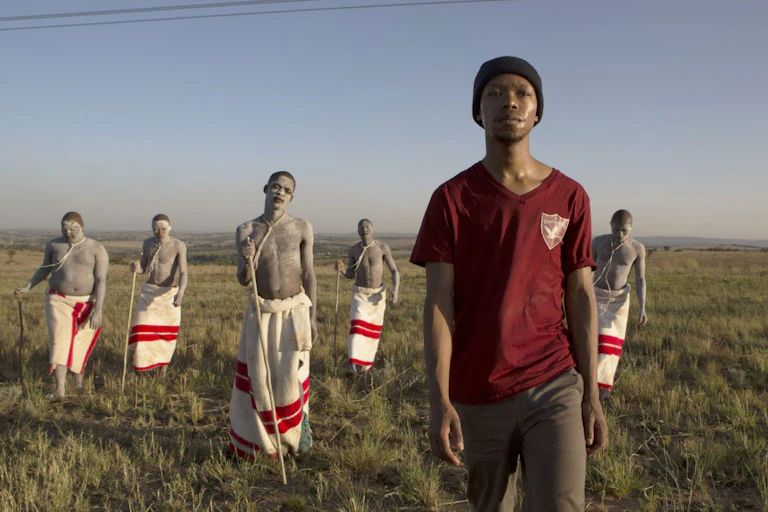 Across an open field, a young man in civilian clothing walks ahead of a group of boys wearing traditional red and white cloths around their waists.