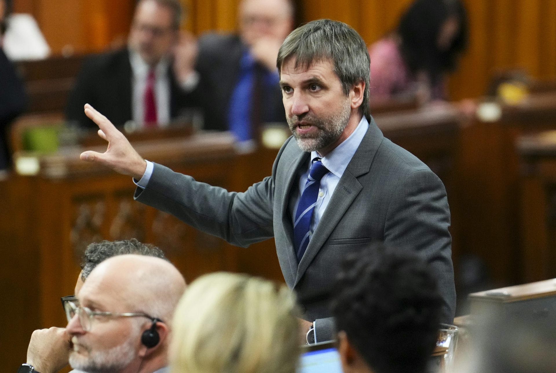 A man wearing a grey suit jacket and tie gestures while speaking to a room full of people.