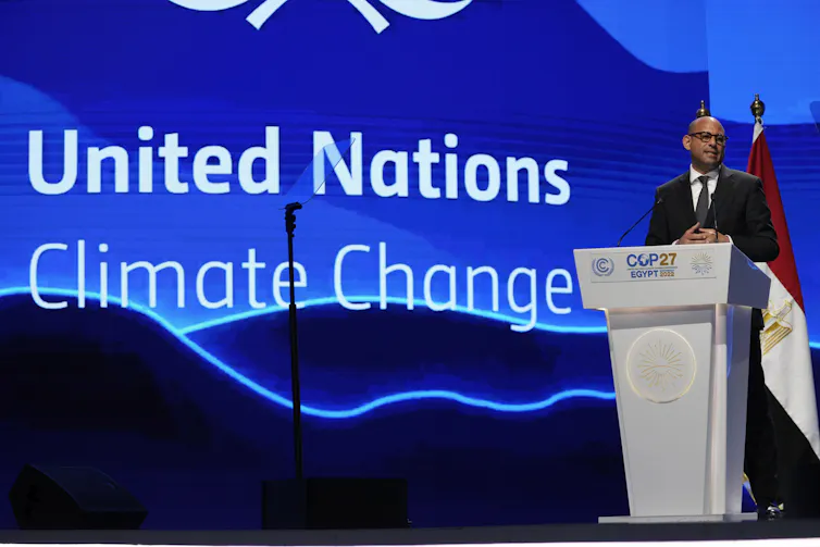 A bald man wearing glasses and a dark suit speaks from behind a podium. Behind him, a screen says 'United Nations Climate Change.