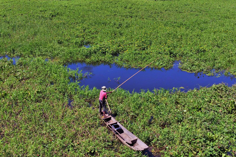 A man with a hat standing in a canoe, handling a fishing rod in a marshy landscape