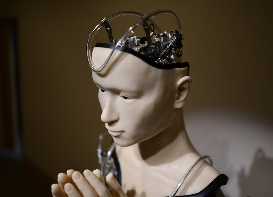 A photograph of a robot with folded hands, showing mechanized equipment in a partial view of the head.