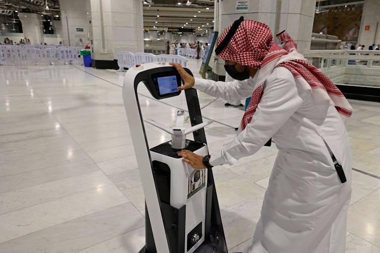 A man in red checked head scarf and flowing white shirt with a robot.