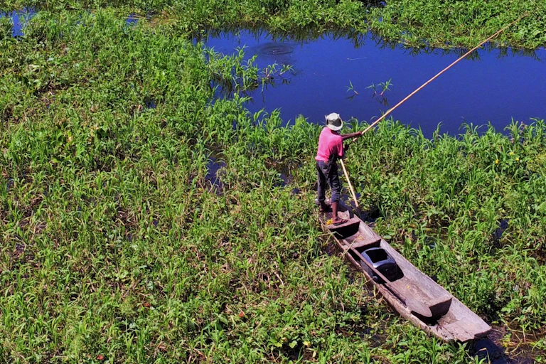 Um homem de chapéu em pé em uma canoa, manuseando uma vara de pescar em uma paisagem pantanosa
