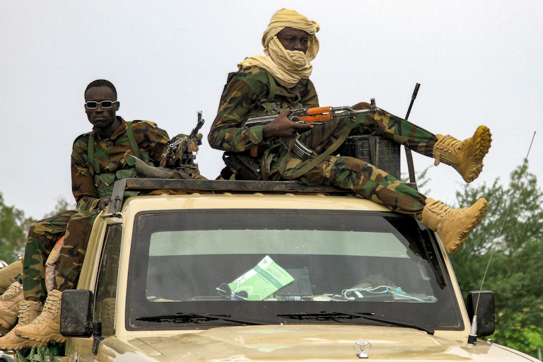 Two armed Black men wearing camouflage sit on the roof of a military vehicle.