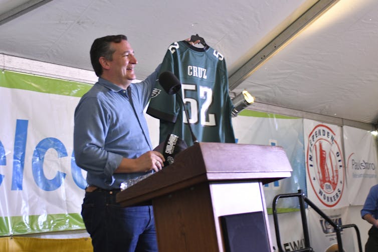 Ted Cruz holds up a green jersey with his name on it while standing at a podium.