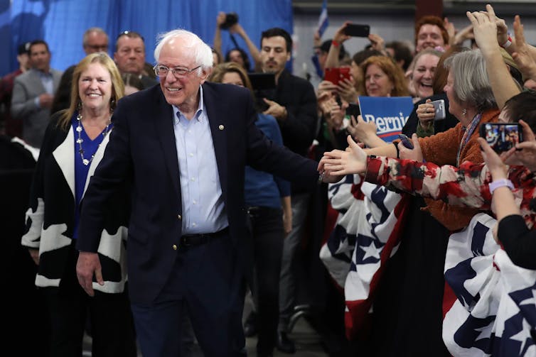 Bernie Sanders walks through a crowd of people smiling, standing in front of his wife.