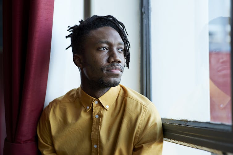 A Black man looks hopeful as he sits near a window and looks outside.